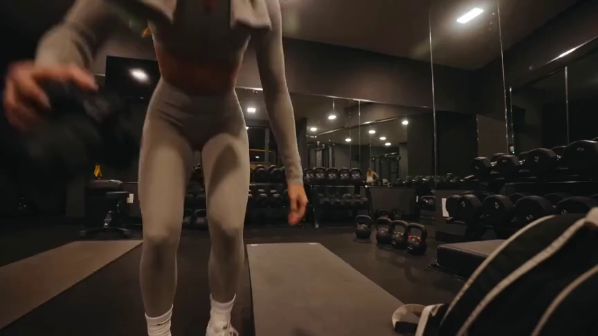 Create a 15-second, 16:9 intimate end-of-workout vlog starring one consistent fictional 24-year-old Brazilian fitness creator, CHASE: slim athletic build, real glowing skin, expressive brown eyes, and long wavy chestnut-brown hair in a loose ponytail. She wears a fitted long-sleeve workout top, high-waisted leggings, white socks, sneakers, and a towel over her shoulders. Handheld DV-camcorder vlog footage with warm 16mm-inspired analog tape: CHASE films herself at arm’s length, then sets the camera on a bench, gym bag, or beside a stretching mat; the camera never appears in frame or reflection. Include subtle shake, drifting framing, autofocus hunting, rushed reframes, uneven zooms, occasional face crops, tape grain, soft lamp halos, exposure shifts, low contrast, and accurate skin. In a mostly empty boutique gym late evening with warm lights, darker corners, mats, mirrors, dumbbells, workout bag, and protein shake, keep her tired, funny, unpolished, breathing audibly, relaxed, and natural. Sequence: on the mat, “Okay… that session is officially over.” Stretching, “I definitely pushed that last set too far.” Shoulder roll, “Tomorrow is going to be interesting.” Close-up sipping a shake, “Wait… this one is actually good.” Side lean, “I’d call today a solid eight and a half.” Resting on one knee, “Exhausted, but in the best way.” Finally bags up and waves: “I’m calling it. See you at the next workout.” Natural English lip sync, blinking, micro-expressions, identical face, hair, wardrobe, and physique. Avoid polished commercial styling, plastic skin, robotic movement, face swaps, added people, captions, logos, watermarks, visible camera, or AI artifacts.